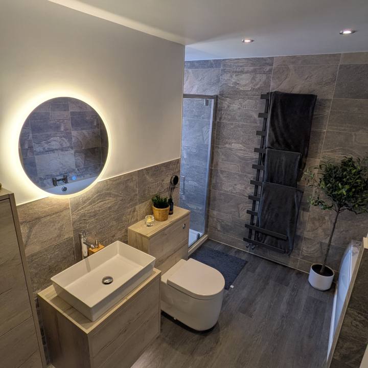 Finished bathroom viewed from the doorway, with grey stone-effect tiles, oak-toned furniture, a round mirror and plants.