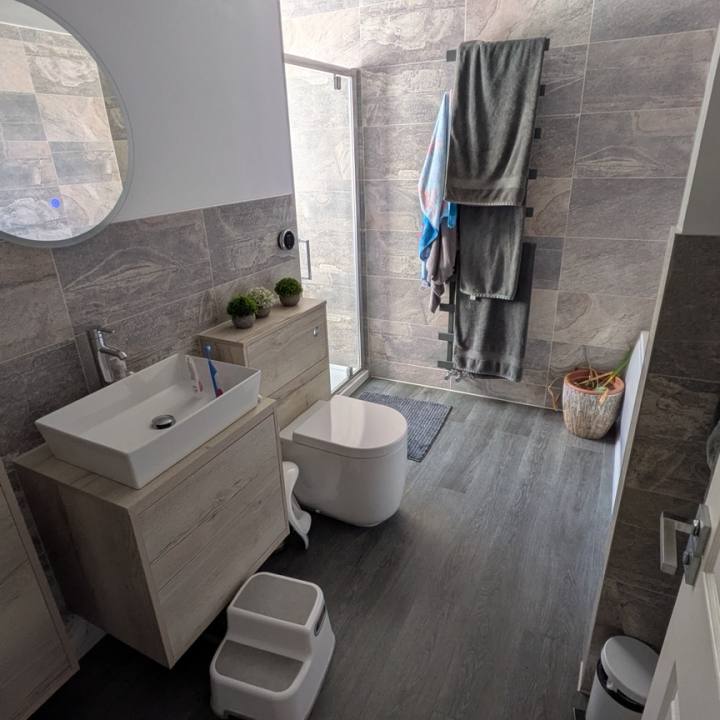 Bathroom before the finishing touches, viewed from the doorway with grey stone-effect tiles, oak-toned furniture and a round mirror.