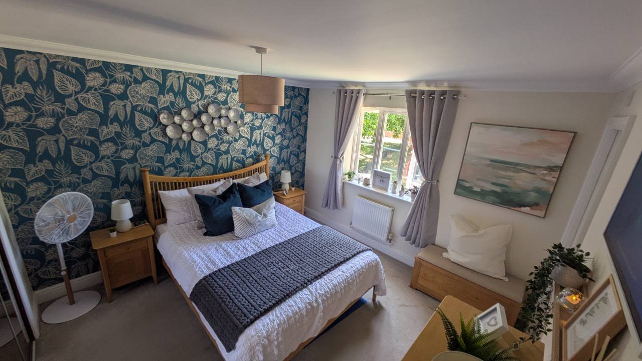 Finished bedroom with teal botanical wallpaper, layered white bedding, soft grey curtains, oak furniture and framed artwork.