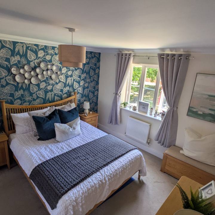 Finished bedroom with teal botanical wallpaper, layered white bedding, soft grey curtains, oak furniture and framed artwork.